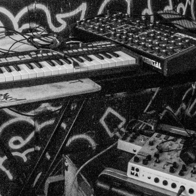 Close-up of an electronic music setup, including a small MIDI keyboard, a synthesizer with multiple knobs labeled "Sequential," and various audio cables and effects units against a graffiti-style background.